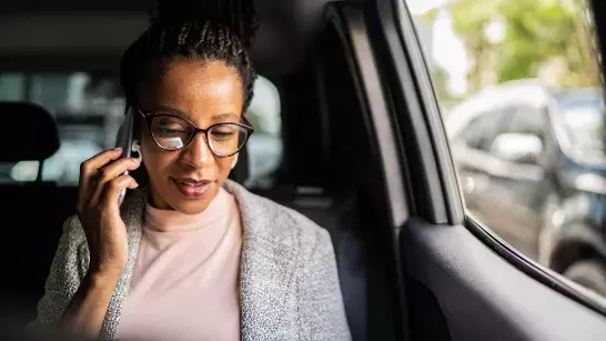 Woman wearing glasses , a grey sweater and a pink tshirt sitting in a car with the window rolled down with a phone held up to her ear