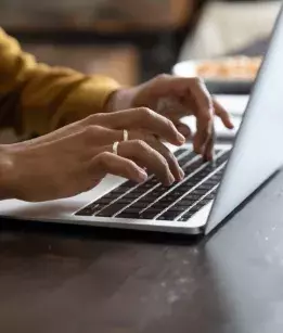 Two hands typing on a laptop keyboard on a brown tabletop. One hand has two silver rings on two fingers