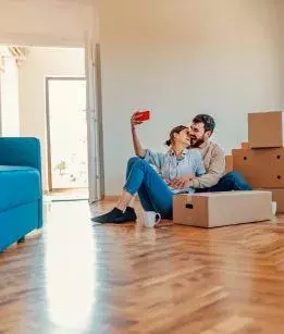 Hew homeowner couple sits on the floor of their living room o take a selfie between boxes