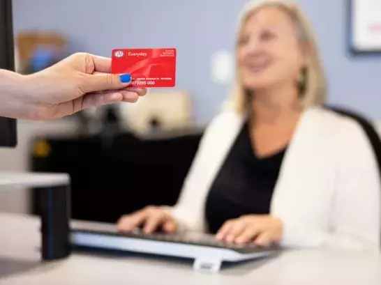 Hand with blue nailpolish holding a red CAA membership card with a woman typing at a computer in the background