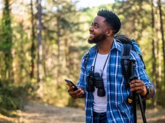 Male holding a phone anda. walking stick wearing a blue plaid shirt over a white shirt, a backpack and binoculars slung over his neck with trees in the background