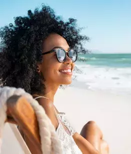 a young afriacn-american woman smiling wearing sunglasses sitting in a beach chair with the ocean in the background