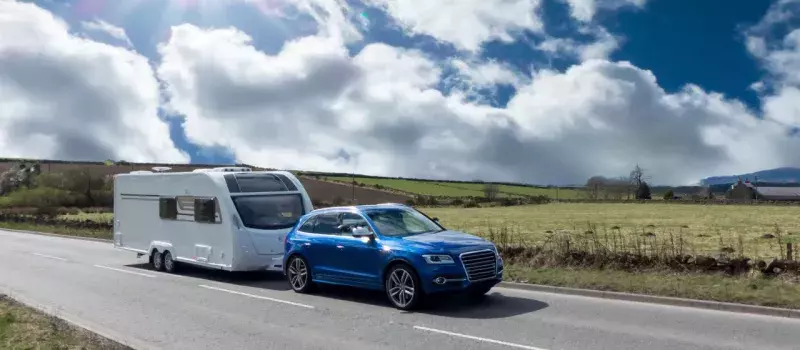 Blue SUV is towing a family camper down a highway surrounded by grassy fields on a sunny day. 