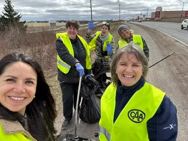 Team of CAA employees in safety vests pick up garbage on a busy road.