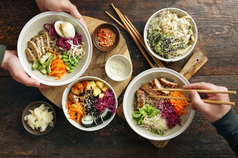 Four colorful bowls of assorted poke are displayed on a wooden table. Ingredients include vegetables, seafood, and rice, with toppings like sesame seeds and avocado. Two pairs of hands with chopsticks reach for food from the bowls. Small side dishes are also present.