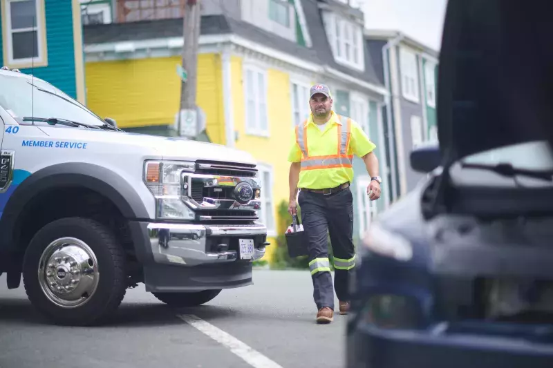 A roadside assistance worker in a high-visibility yellow shirt and cap walks toward a vehicle with an open hood. A service truck with "Member Service" written on the door is parked nearby. Colorful houses line the street in the background.