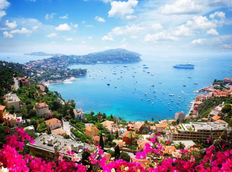 A vibrant coastal view with turquoise waters, numerous boats, colorful flowers, and red-roofed houses along the hillside.