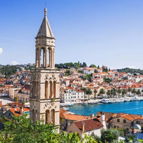 A tall stone bell tower rises above a coastal town with orange-tiled rooftops, palm trees, and a marina along clear blue water.