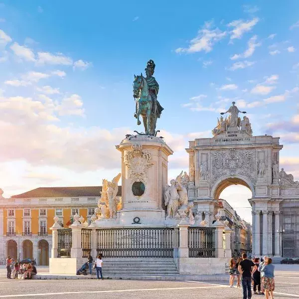 A grand equestrian statue stands in the center of a large, open plaza with a decorative archway in the background. The surrounding buildings feature bright yellow façades, and people stroll around under a clear, sunny sky.