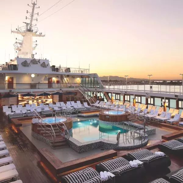 A cruise ship’s open-air deck at sunset, featuring multiple small pools and hot tubs in the center, surrounded by striped lounge chairs. The ship’s mast and upper decks rise in the background, with a distant shoreline silhouetted against a warm orange sky.