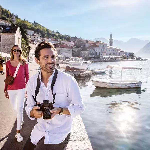 A man with a camera and a woman walk along a scenic European waterfront with boats, stone buildings, and mountains.