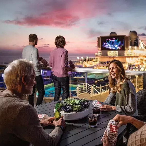 People on a cruise ship deck at sunset, enjoying drinks, playing cards, and watching entertainment on a large outdoor screen.