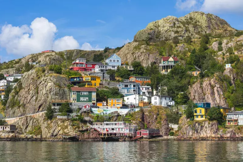 A picturesque coastal village with colorful houses built on rocky cliffs. The houses are painted in various bright colors such as red, blue, yellow, green, and white. The village is situated on a hillside with lush greenery and trees scattered throughout. The sky is clear with some fluffy white clouds, and the calm water at the bottom reflects the vibrant buildings above.