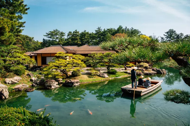 A serene Japanese garden with a pond, lush greenery, and a traditional building in the background. Two people are in a small wooden boat on the pond, one standing and rowing with a long pole while the other is seated. Colorful koi fish swim near the surface of the pond, surrounded by well-manicured trees and shrubs under clear blue skies.