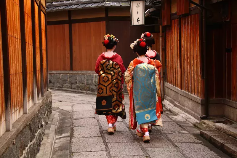 Three individuals dressed in traditional Japanese kimonos walking down a narrow, stone-paved street lined with wooden buildings. They have elaborate hairstyles adorned with hair accessories and are wearing wooden sandals. A sign with Japanese characters hangs above the street.