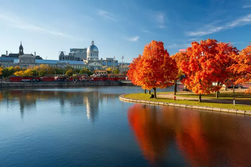 A serene waterfront scene with vibrant autumn foliage. Bright orange and red trees reflect in the calm water, with a historic building featuring a dome and other architectural structures in the background. The sky is clear with some wispy clouds.