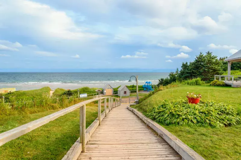 A wooden boardwalk leads down to a beach, flanked by grassy areas and a red flower pot. The ocean and waves are visible under a partly cloudy sky, with small structures like a shed and possible playground equipment near the shore. Trees and shrubs line the right side.