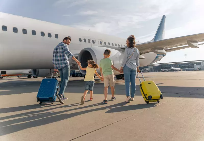 A family of four walks toward a white airplane on the tarmac, holding hands and pulling blue and yellow suitcases.