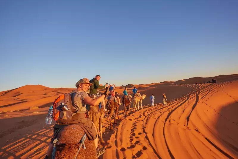 People ride camels through a desert with sand dunes under a clear blue sky, leaving tracks and shadows in the sand.