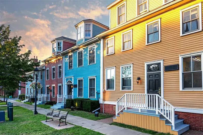 A row of red, blue, and yellow townhouses with porches, set along a sidewalk with grass, a bench, and street lamps.