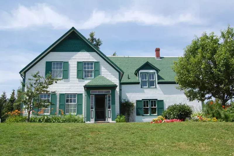 A white house with green trim, shutters, and a gabled roof sits among trees, bushes, and flowers under a partly cloudy sky. This is the Green Gables Heritage Place in Prince Edward Island.