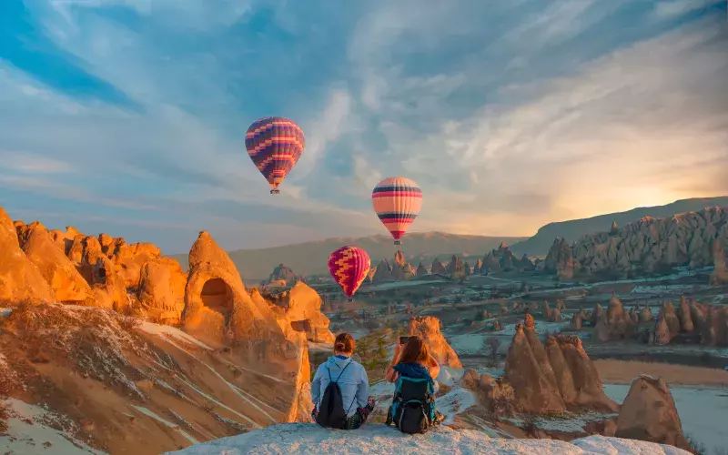 Two people sit on a rock ledge watching three hot air balloons float over a rocky landscape at sunrise or sunset.