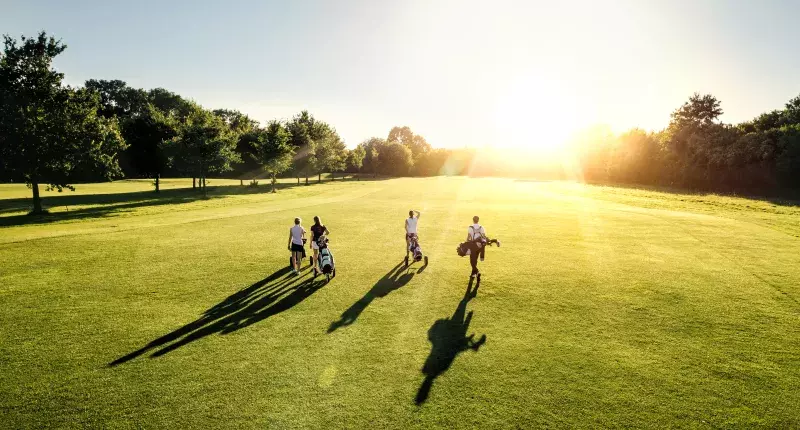 Four people walking on a golf course at sunset, carrying golf bags and pushing golf carts.