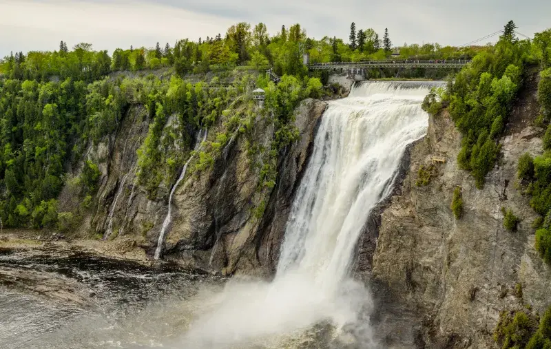 A large waterfall flows down a rocky cliff with a bridge above and lush greenery surrounding it.