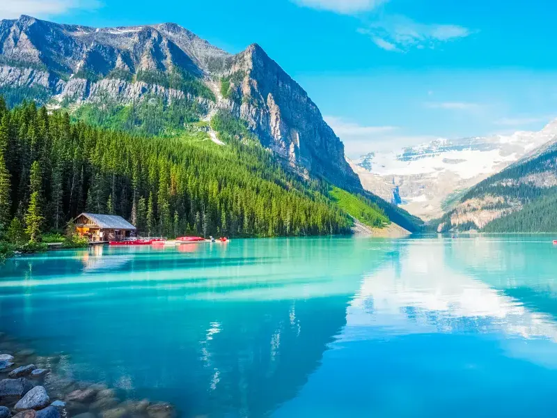 A turquoise mountain lake with a wooden cabin and red canoes, surrounded by forests and snow-capped peaks.