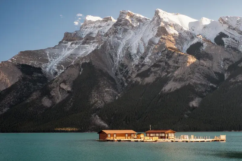 Two wooden cabins on a floating dock over turquoise water, with snow-capped mountains in the background.