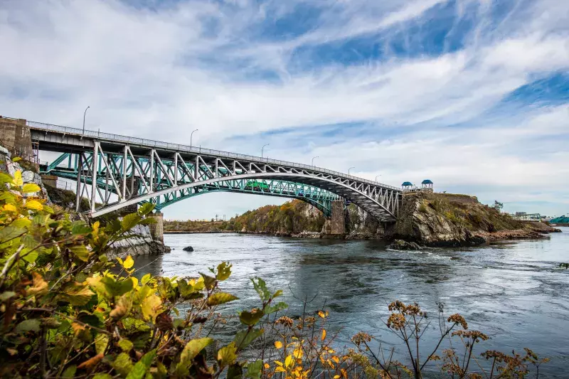 Reversing Falls Bridge in Saint John, spanning water with autumn foliage in the foreground and a partly cloudy sky.