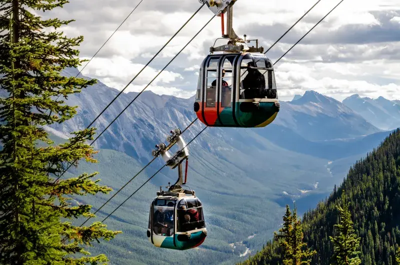 Two gondola cabins travel through a mountainous landscape with rugged peaks and dense forests under a partly cloudy sky.