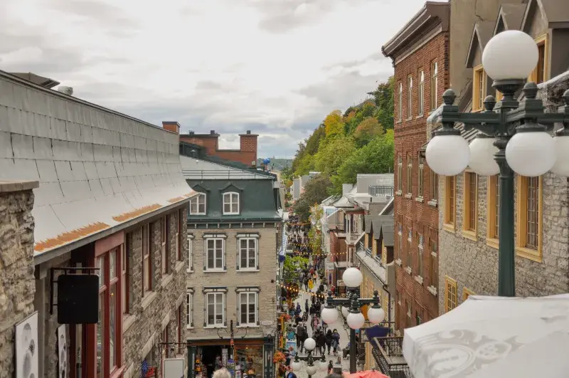 A busy old town street lined with historic stone buildings and street lamps under a cloudy sky.