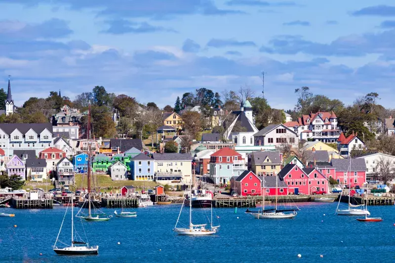 Colorful waterfront buildings and boats in Lunenburg, Nova Scotia, with hillside houses, a white church steeple, and a partly cloudy sky.