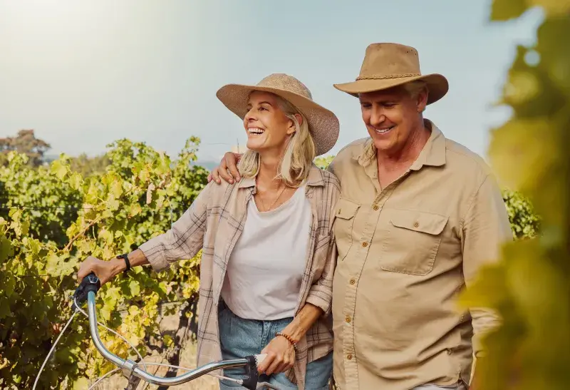 Two people standing together in a vineyard, wearing hats and casual outdoor clothing, with one person’s arm around the other. Lush green grapevines and a clear sky form the background.