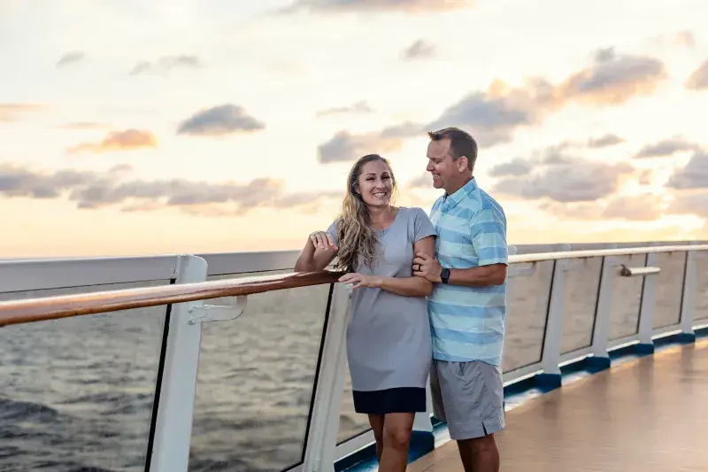 Two people stand on the deck of a ship, gazing at the ocean during a vibrant sunset.
