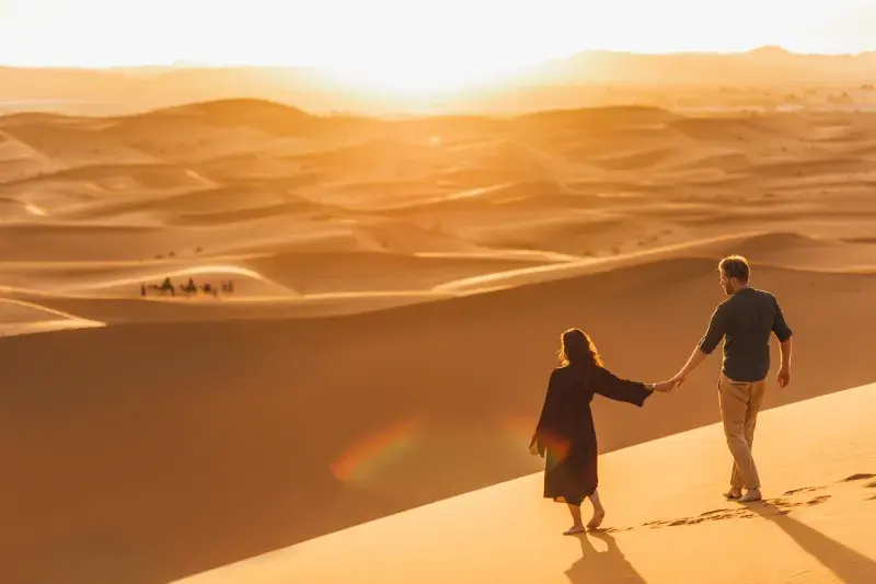 Two people walk hand-in-hand across expansive sand dunes during a golden sunset or sunrise.