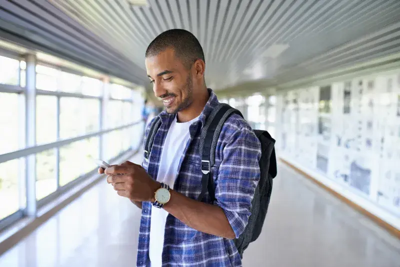 Man in plaid shirt with backpack looking at phone in a bright hallway.