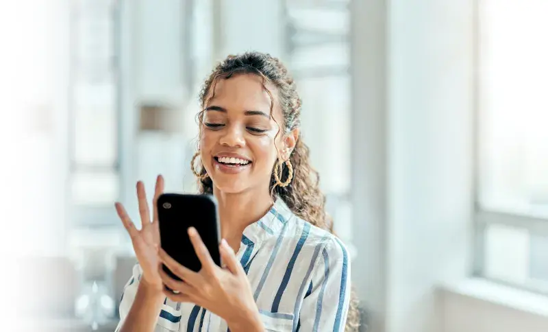 Person in striped shirt smiling at smartphone in a sunlit indoor space.