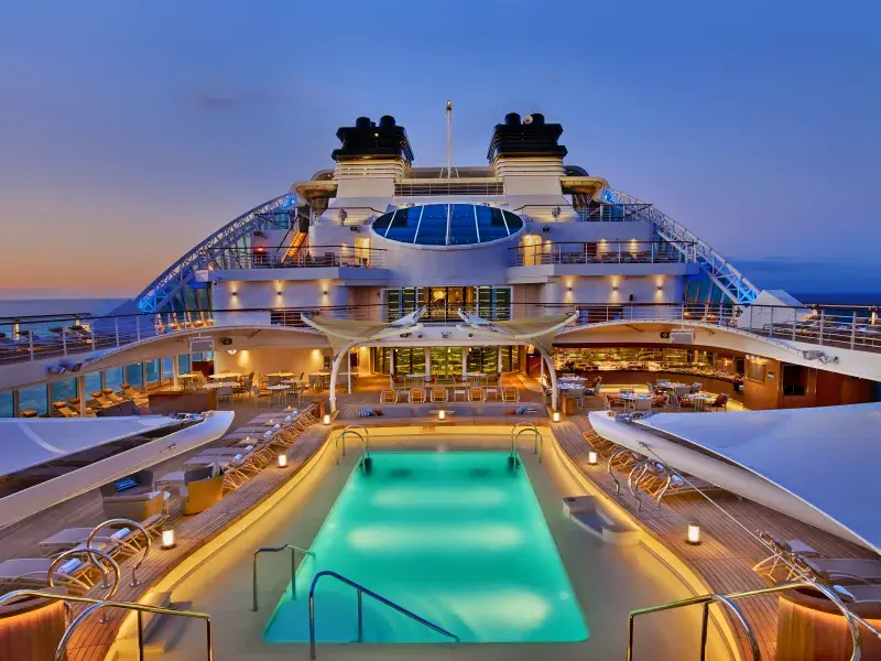 Cruise ship deck at dusk with a turquoise-lit pool, lounge chairs, and upper-level dining areas under a twilight sky.