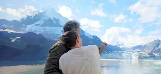 Two people standing on a cruise ship balcony, embracing while looking out at snow-capped mountains and a glacier under a bright blue sky.