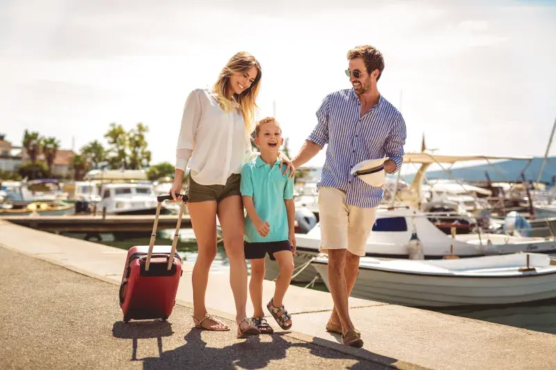 Family walking along a marina with rolling luggage, boats docked in the background.