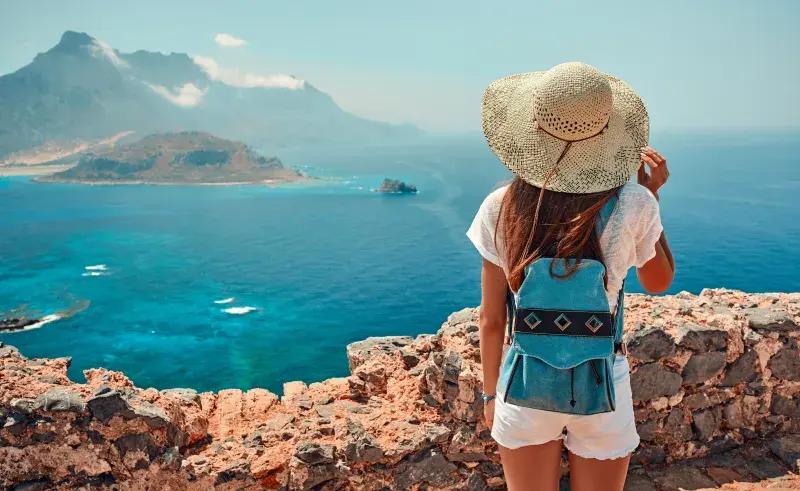 A person wearing a sunhat, white shirt, and a small blue backpack stands on a rocky viewpoint overlooking a bright blue sea with distant mountains and a small island in the background.
