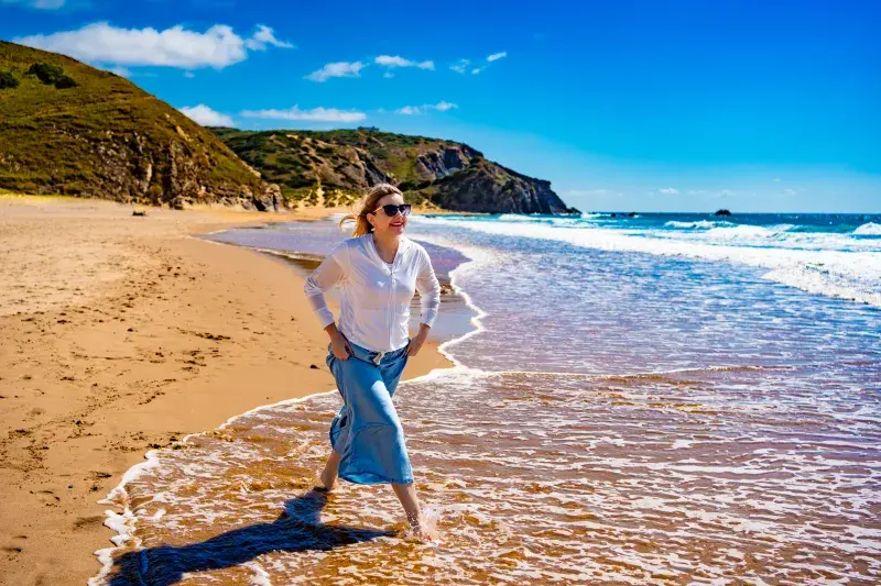 Person walking barefoot along a sandy beach shoreline, with waves, cliffs, and a bright blue sky.