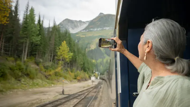 Woman on a train holds up a phone to photograph a forested mountain landscape.