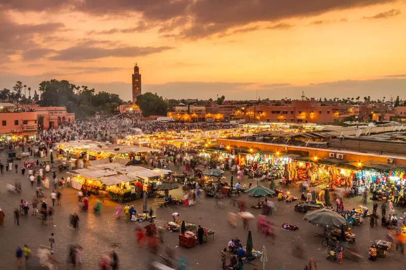 Busy market square in Marrakech at sunset, filled with glowing stalls, crowds of people, and the Koutoubia Mosque rising in the background.
