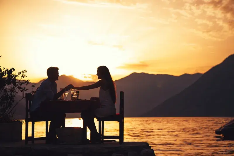 Two people seated at a small table by the water at sunset, facing each other with glasses on the table. The scene is backlit by a golden sky, with mountains in the background and the water reflecting the warm light.