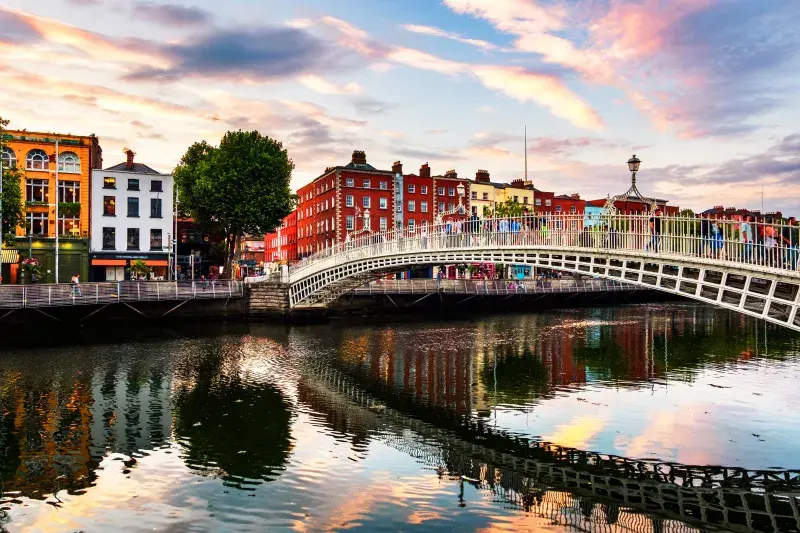 The Ha'penny Bridge spanning the River Liffey in Dublin at sunset, with colourful historic buildings lining the waterfront and reflections shimmering on the water.