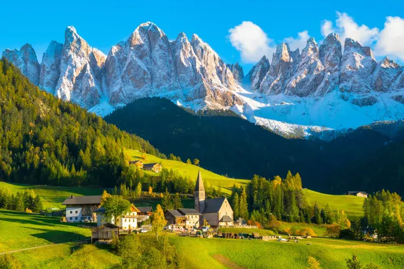 Snow-capped peaks of the Dolomites rise above a green alpine valley with forested hills, a small village, and a church surrounded by sunlit meadows under a clear blue sky.