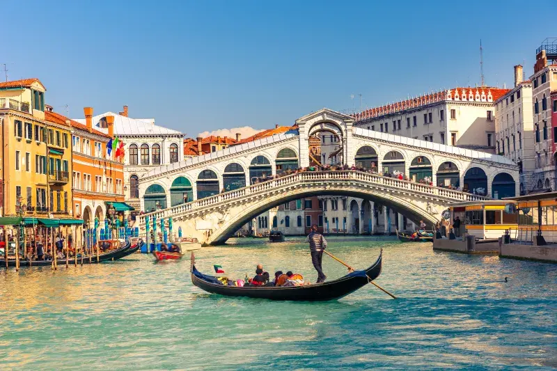 A gondola travels along Venice's Grand Canal in front of the historic Rialto Bridge, with colourful buildings, boats, and waterfront activity lining the canal on a clear day.